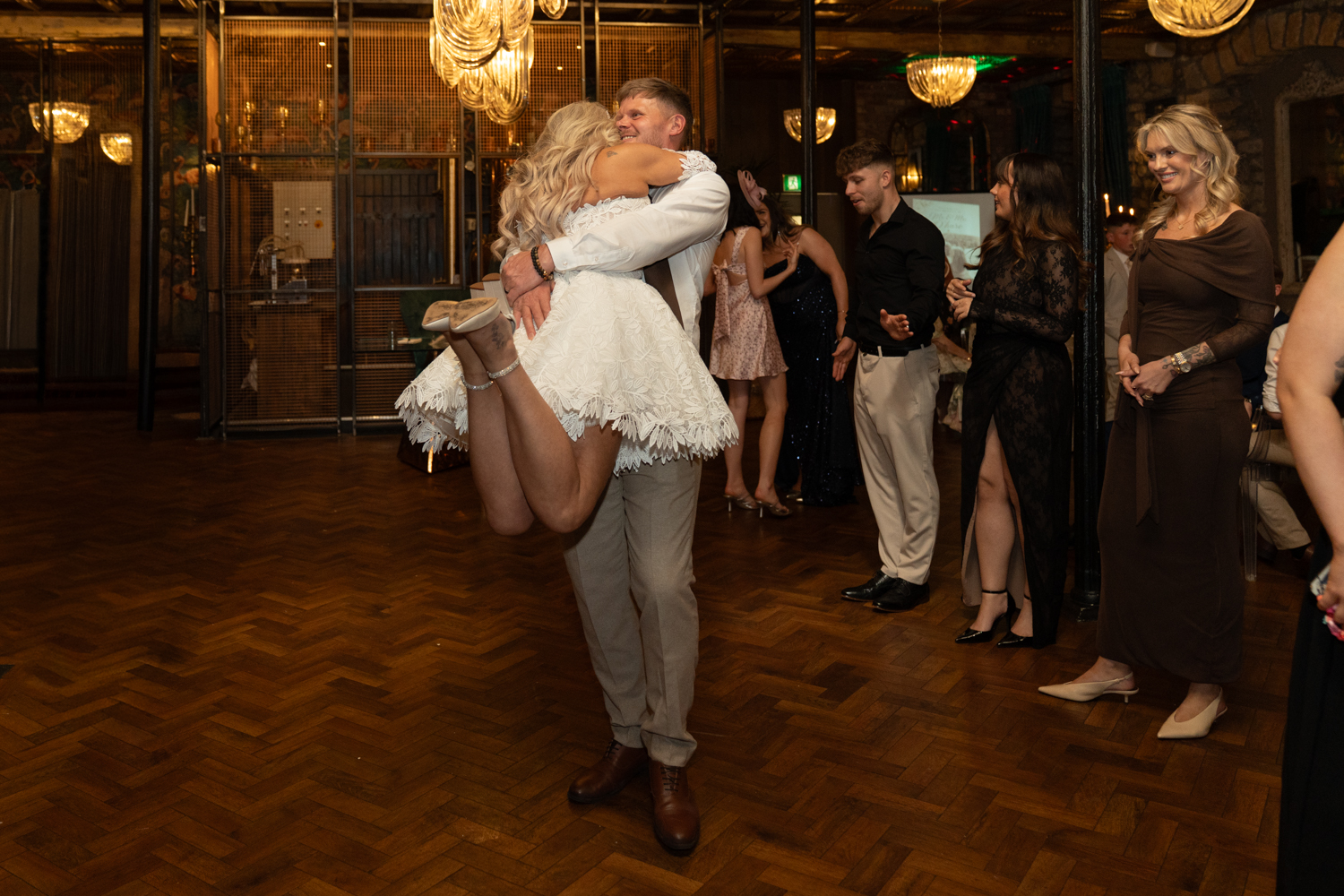 The groom lifts the bride off her feet during their first dance — her arms around him, guests smiling in the background